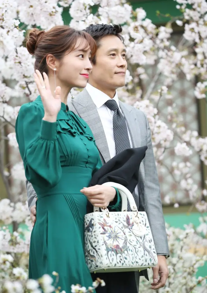A woman in a green dress waves while posing with a man in a gray suit, set against a backdrop of blooming cherry blossoms.