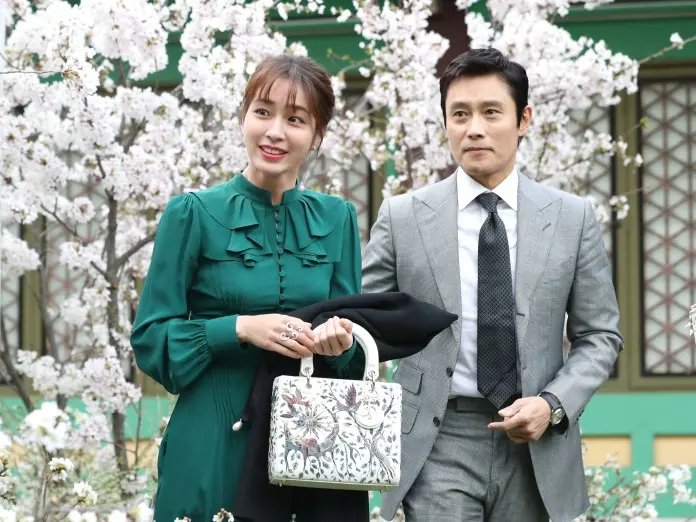 A woman in a green dress and a man in a gray suit are smiling as they walk together among blooming cherry blossom trees.