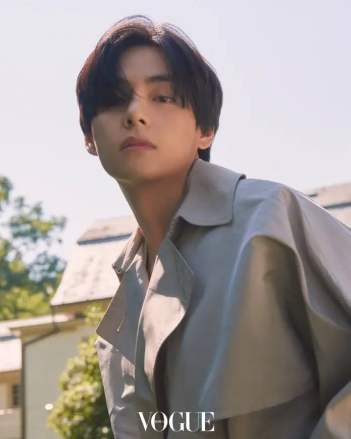 V with long dark hair is looking thoughtfully at the camera while wearing a stylish beige trench coat, set against a sunny outdoor background.