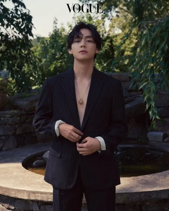 V wearing a pinstripe suit stands confidently outdoors, surrounded by greenery, with a natural stone fountain in the background.