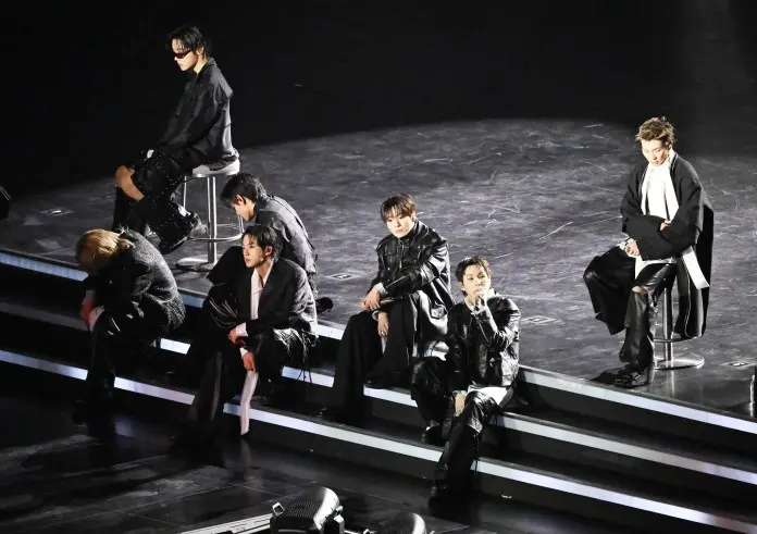 BTS dressed in black outfits seated on steps during a concert, with a spotlight illuminating them from above.