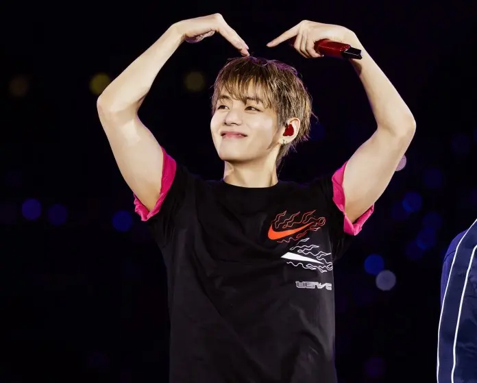 V smiling and making a heart shape with his hands, wearing a black t-shirt with pink accents, on stage with a colorful background.