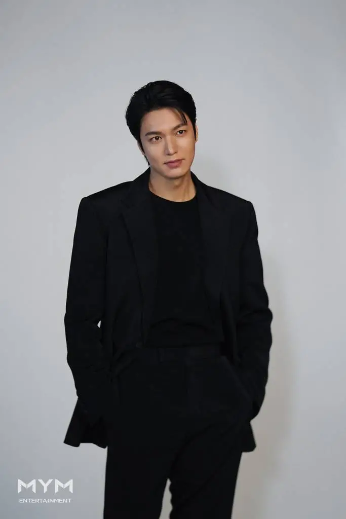 Lee Min-ho in a black suit stands confidently against a neutral background, looking slightly to the side with a subtle smile.