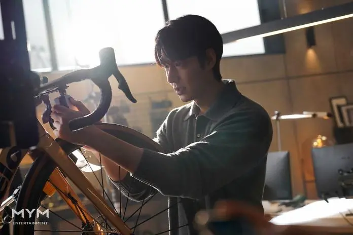 Lee Min-ho inspecting and adjusting a bicycle in a well-lit workspace, with desktop computers and office items in the background.