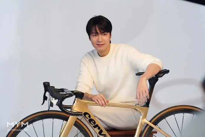 Lee Min-ho dressed in a white sweater and pants, sitting on a stool beside a gold road bicycle, smiling at the camera.