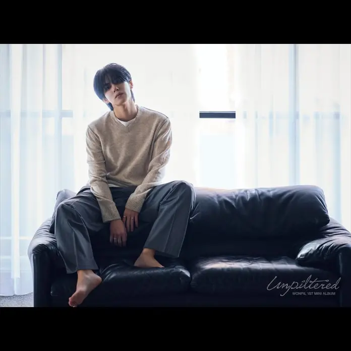 Wonpil sitting casually on a black leather couch, wearing a light sweater and gray pants, with bare feet. Soft natural light filters through sheer curtains in the background.