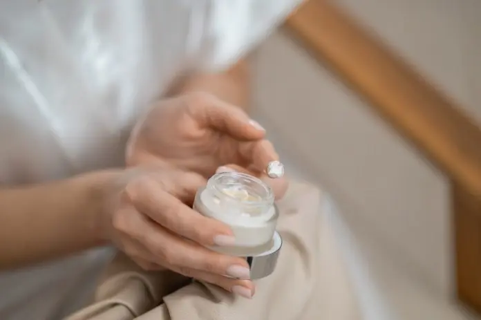 A close-up of a person's hands holding a small jar of cream, with a small amount of product on one fingertip.
