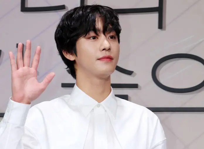 A young man with curly black hair wearing a white shirt is raising his right hand, smiling slightly, against a minimalistic background.