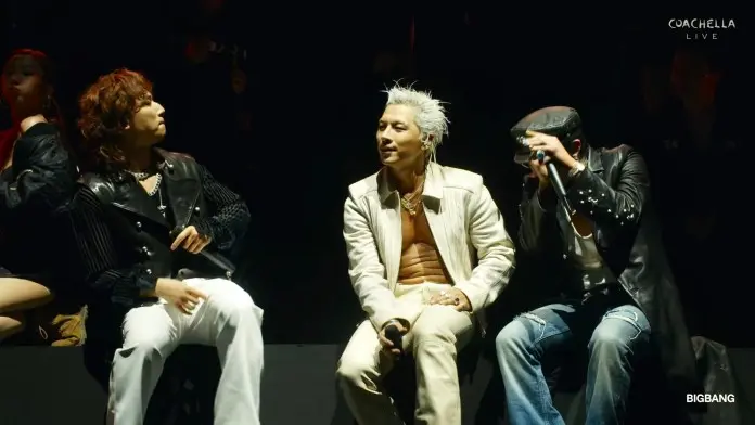 BIGBANG sitting on stage during a live concert, with one wearing a white outfit and two in darker attire, showcasing an energetic atmosphere.