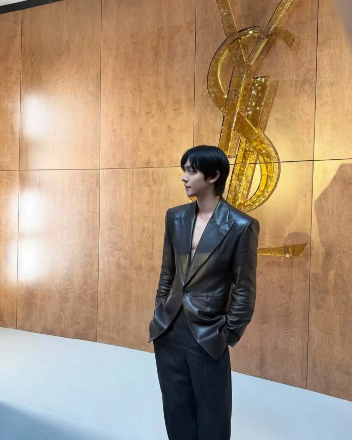 Ahn Hyo-seop beside a large illuminated YSL logo, wearing a stylish black leather blazer and pinstripe pants, against a wooden wall backdrop.