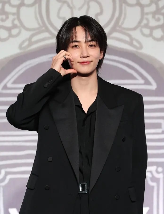Jeonghan in a black suit and shirt is smiling and making a heart gesture with their hand.