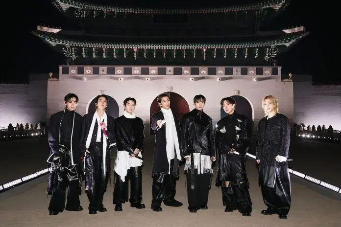 BTS dressed in stylish, modern black outfits stands in front of a traditional architectural structure at night, with soft lighting illuminating the scene.