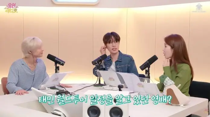 Taemin sitting at a radio studio table with microphones, engaged in conversation. One person has short white hair and wears a blue striped shirt, another has brown hair in a denim jacket, and the third has long brown hair in a green sweater.