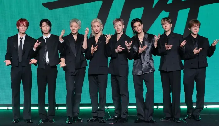A group of eight men in black suits, standing in a row with various poses, against a green screen background.