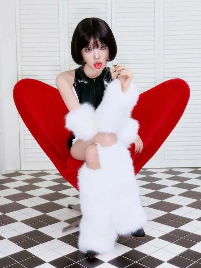 Choi Yena with a bob haircut sitting in a large red heart-shaped chair, wearing a black latex outfit and white fur accessories, playfully holding a red lollipop.