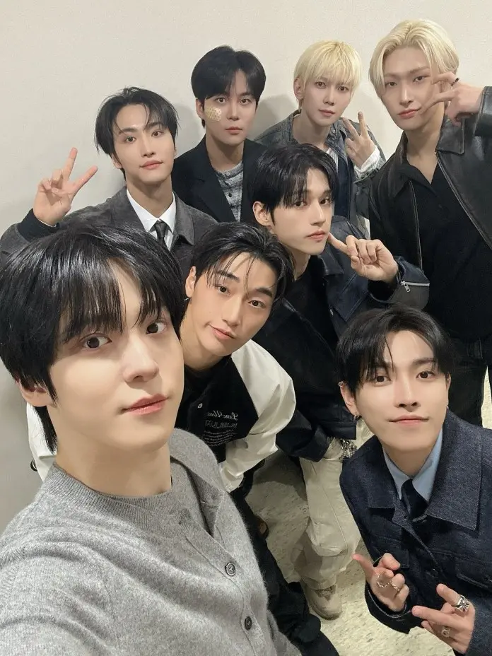 ATEEZ posing together with various expressions and styles, some making peace signs and wearing fashionable outfits in a casual indoor setting.