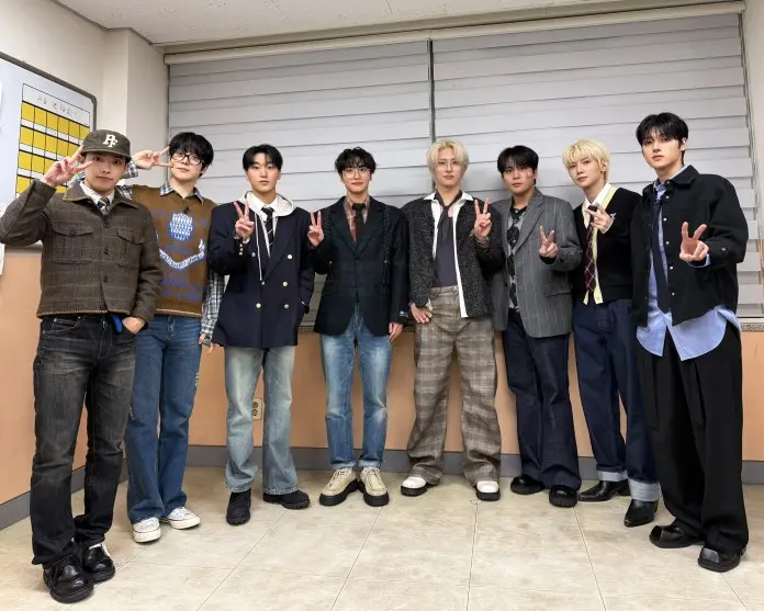 ATEEZ posing for a photo indoors, wearing a mix of casual and semi-formal outfits. They are all making peace signs with their fingers and standing in front of a light-colored wall with a window blind.