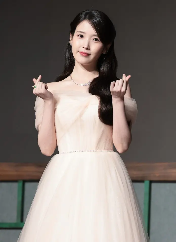 IU wearing a strapless, pale pink gown with a tulle skirt, smiling and making a heart gesture with her hands.