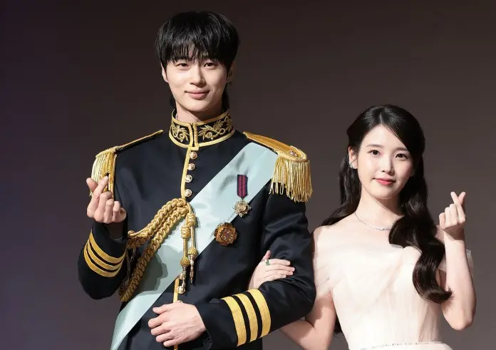  Byeon Woo-seok in a military-style uniform with gold accents and a sash poses next to IU in a light-colored evening gown, both making small heart gestures with their hands against a neutral backdrop.