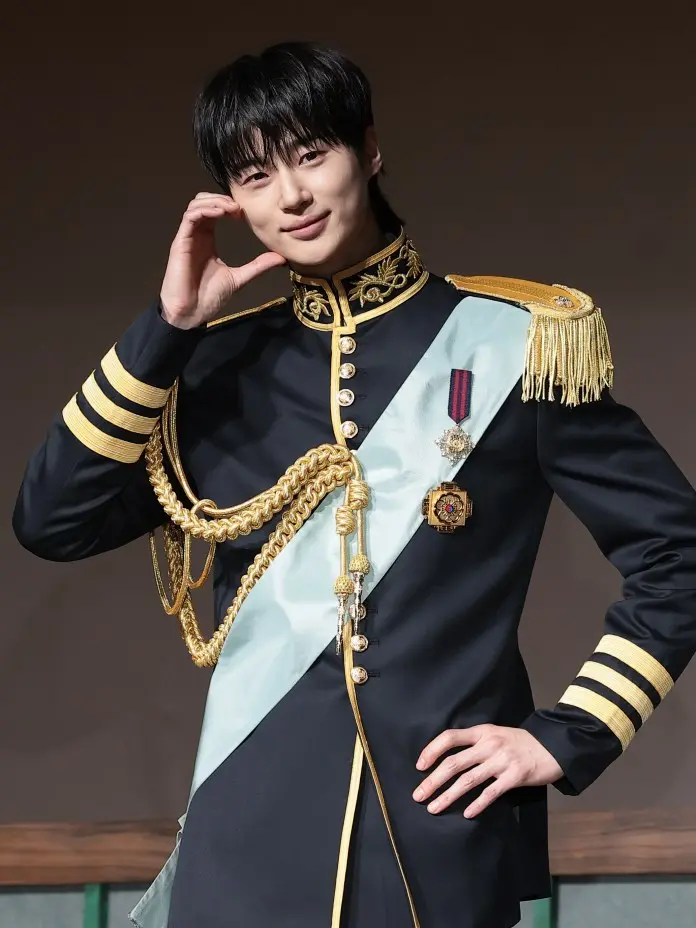  Byeon Woo-seok  in a historical military-style uniform with gold embellishments and a light blue sash, posing with a friendly smile and hand on his chin.