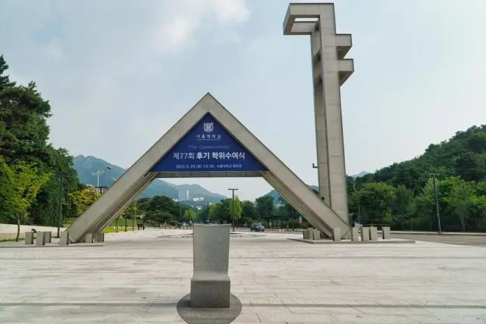 Main entrance archway of a university, featuring a triangular structure with a sign for the 77th commencement ceremony, surrounded by greenery and mountains in the background.