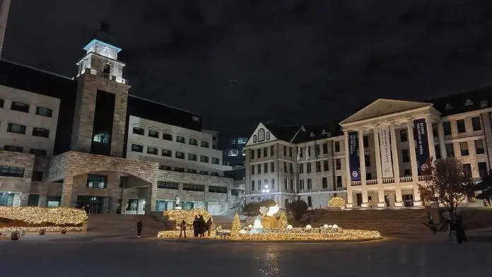 A nighttime view of a university campus featuring modern and classical architecture with decorative lights along the pathways and trees.