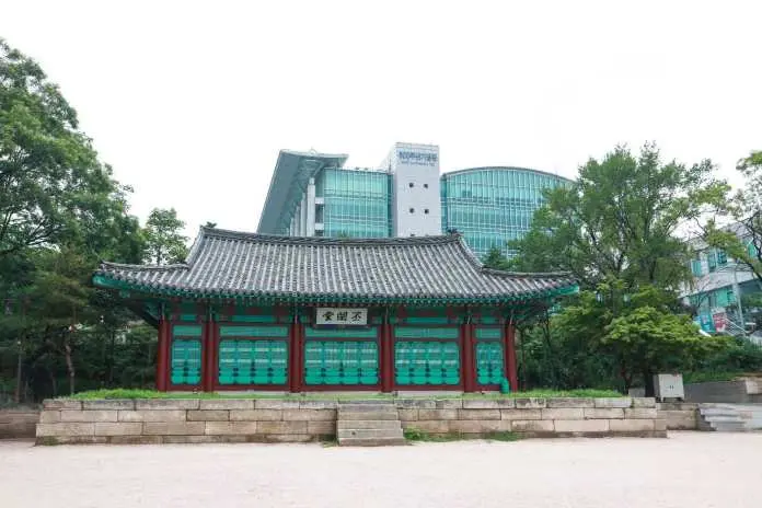 A traditional Korean pavilion with vibrant colors in the foreground, set against a modern building and lush greenery in the background.