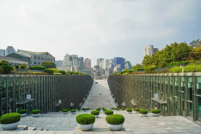 A wide view of a modern urban landscape, featuring a pathway flanked by glass structures and neatly trimmed greenery, leading to a skyline of tall buildings under a partly cloudy sky.