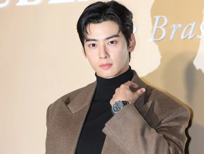 Cha Eun-woo in a stylish brown coat over a black turtleneck, posing confidently with one hand on his shoulder, showcasing a watch.