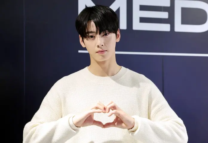 Cha Eun-woo in a cream-colored sweater making a heart shape with his hands, standing in front of a backdrop with the word 'MEDIA.'