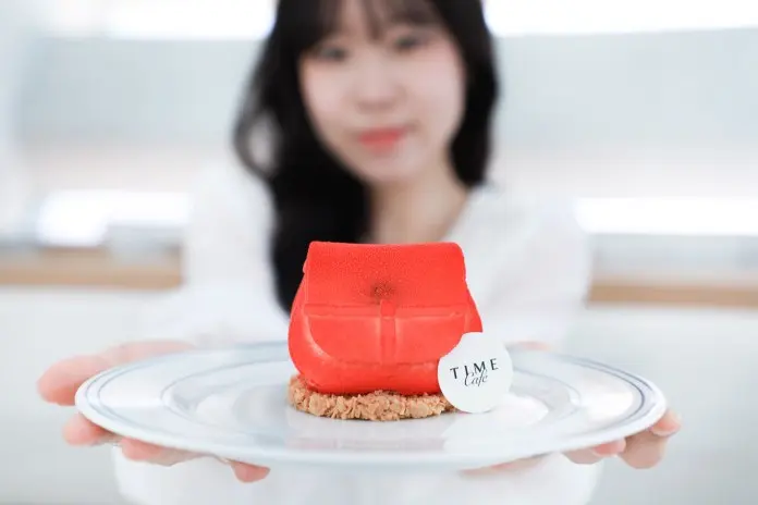 A person holding a plate with a red handbag-shaped dessert, featuring a round tag that reads 'TIME Cafe'. The background is softly blurred, emphasizing the dessert.