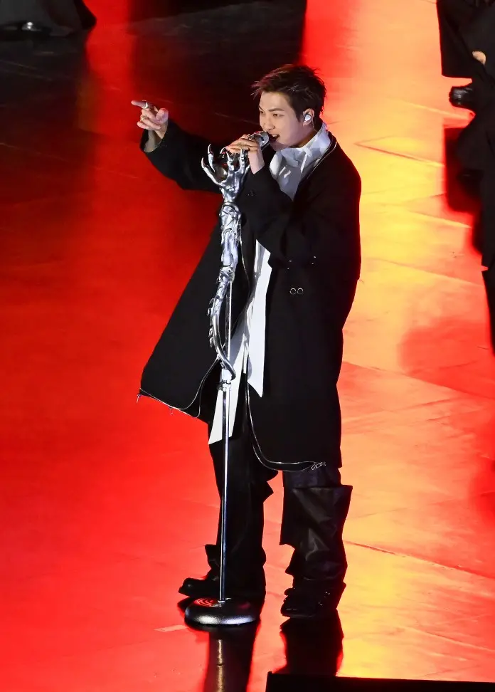 RM on stage singing into a microphone, wearing a black coat and white shirt, with dramatic lighting highlighting the scene.