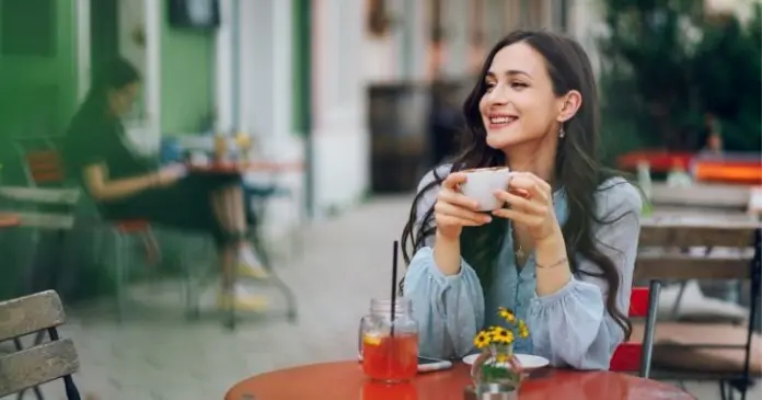 A woman sitting at a table in a café, smiling while holding a cup. A glass with a drink and a small vase with flowers are on the table, with another person in the background.