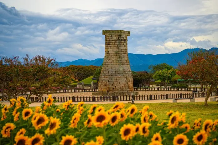 A historic stone tower surrounded by colorful sunflowers and lush greenery, with mountains and clouds in the background.