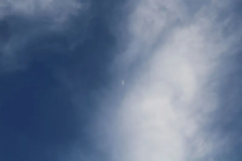 A serene daytime sky with white clouds framing a crescent moon.