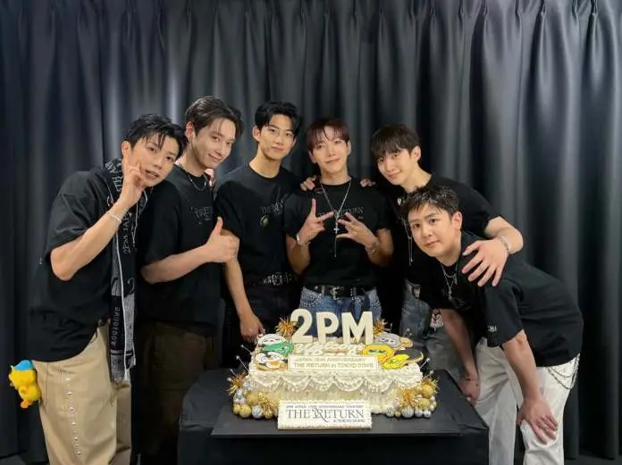 2PM members posing together in front of a decorated cake celebrating the 18th anniversary of a concert. They are all wearing black shirts and smiling, with one member making a peace sign.