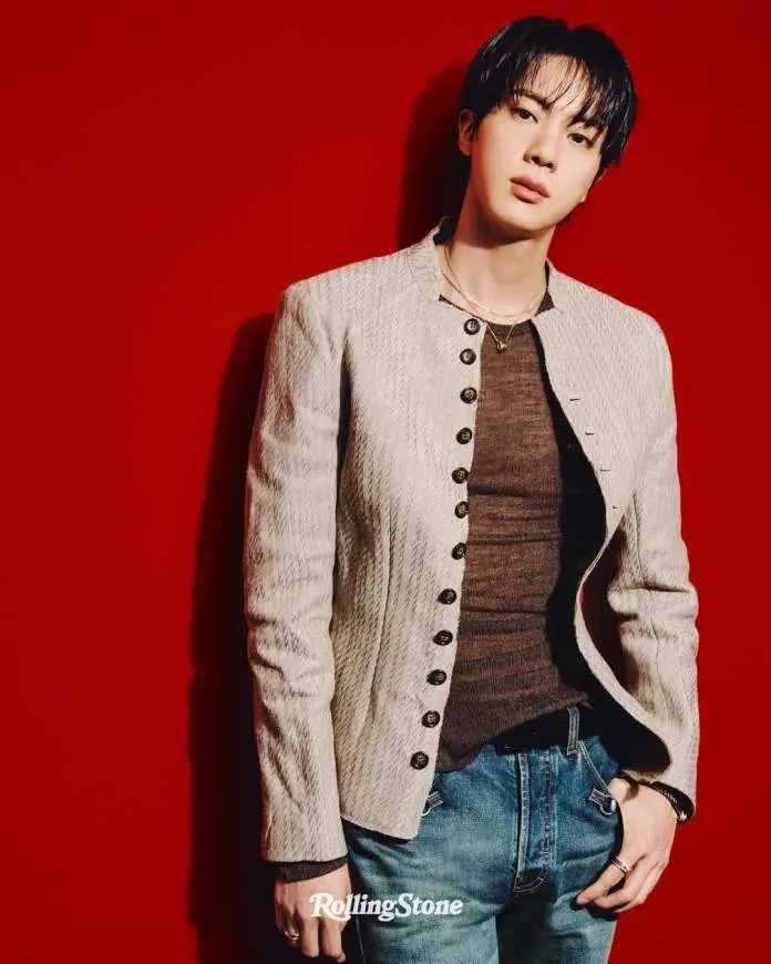 BTS Jin stands against a bright red background, wearing a stylish light jacket over a fitted top and jeans, exuding a confident expression.