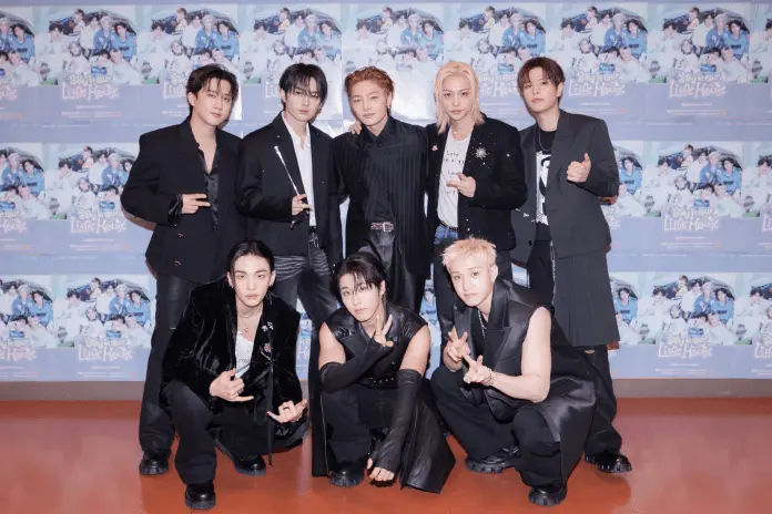 Stray Kids posing for a photo, dressed in stylish black outfits, with a colorful promotional backdrop featuring scenes from a project titled 'Stay in Our Little House'.