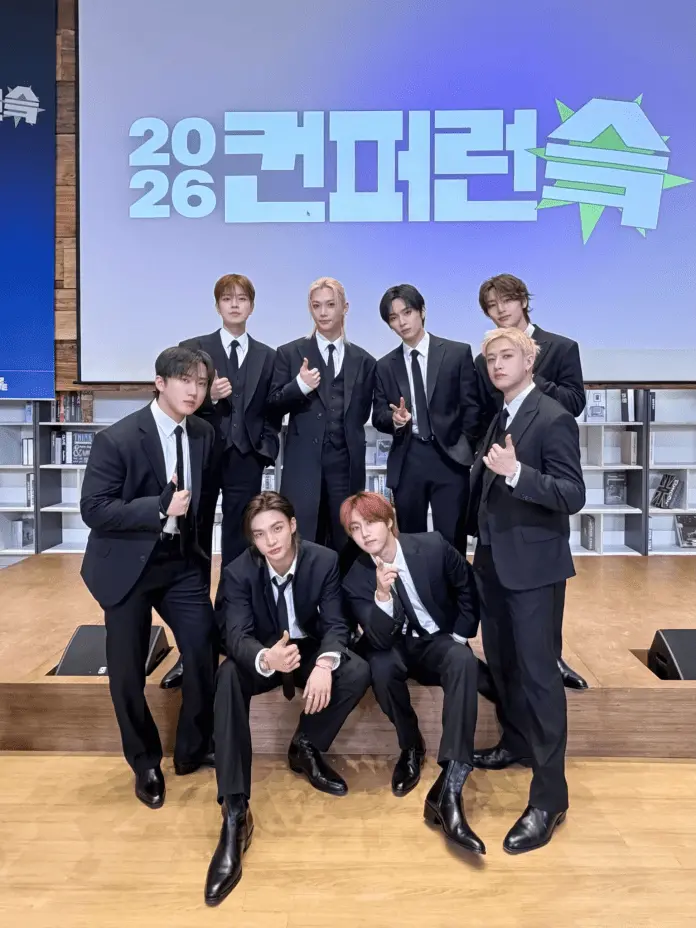 Stray Kids dressed in black suits poses for a photo in front of a backdrop that reads '2026 컨퍼런스'. The group displays gestures such as thumbs up and peace signs, with a mix of standing and seated positions.
