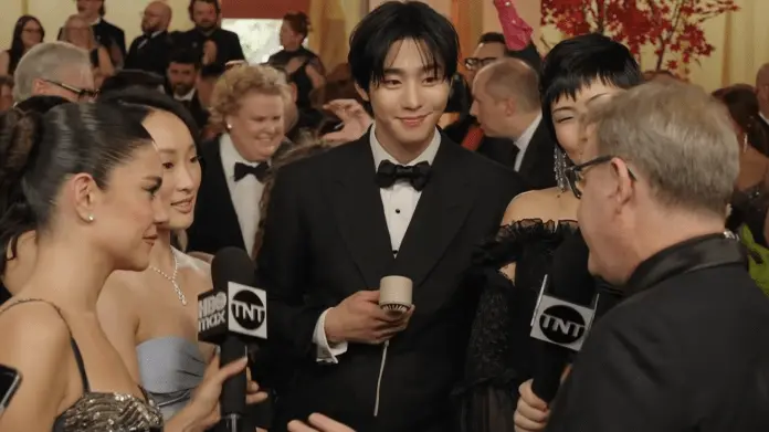 A stylish group of individuals, including Ahn Hyo-seop  in a tuxedo and several women in elegant gowns, engage in conversation during a red carpet event, with cameras and onlookers in the background.