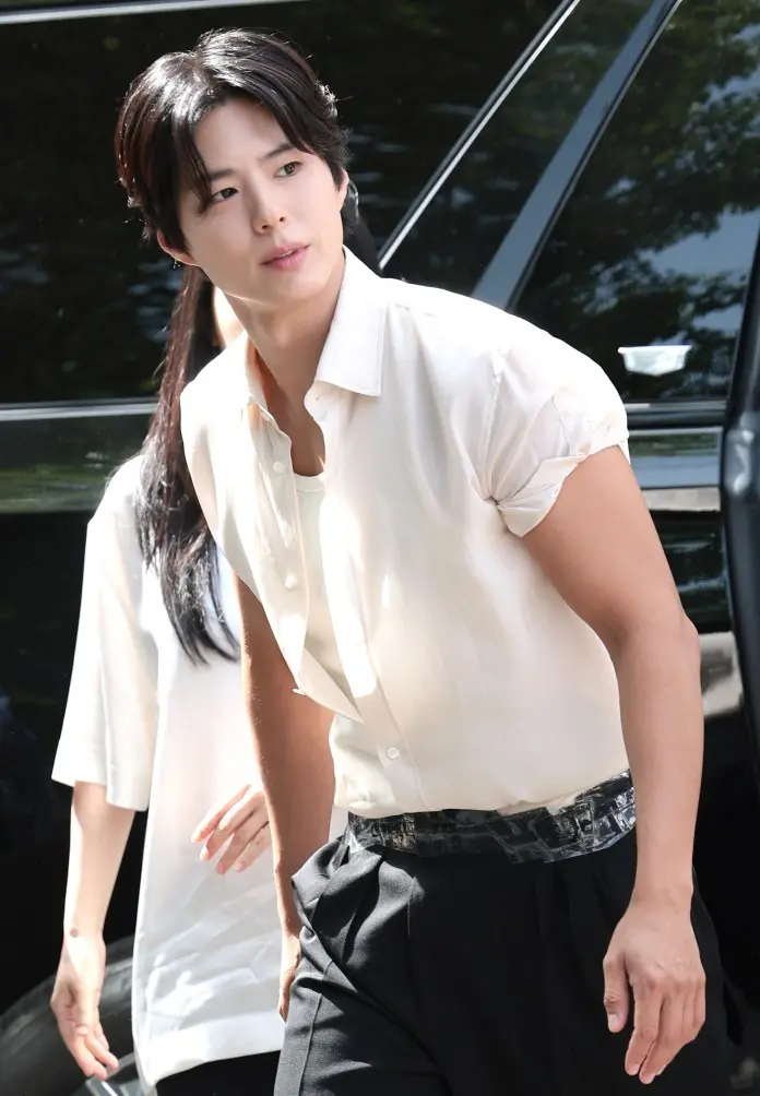 Park Bo-gum with dark hair wearing a white shirt and black trousers walking towards the camera, with a woman in a white top slightly behind him.