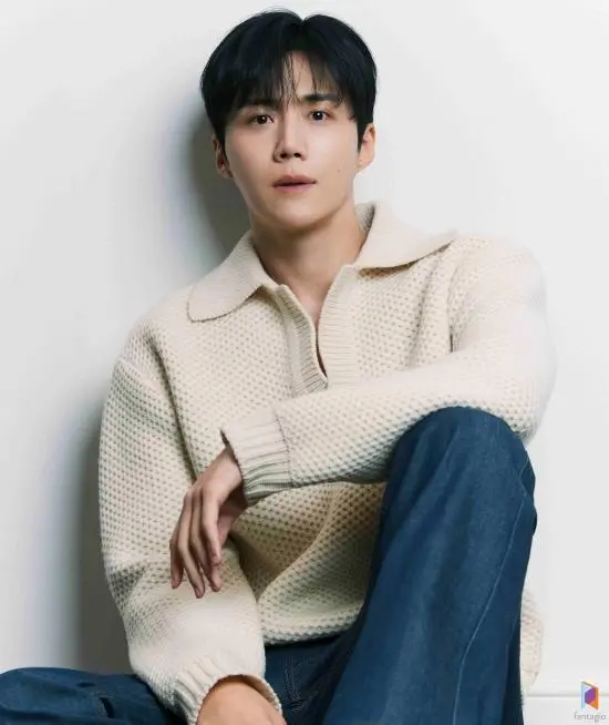 Kim Seon-ho in a light beige knit sweater with a collar, sitting on the floor and looking at the camera, styled with dark hair and wearing blue jeans.