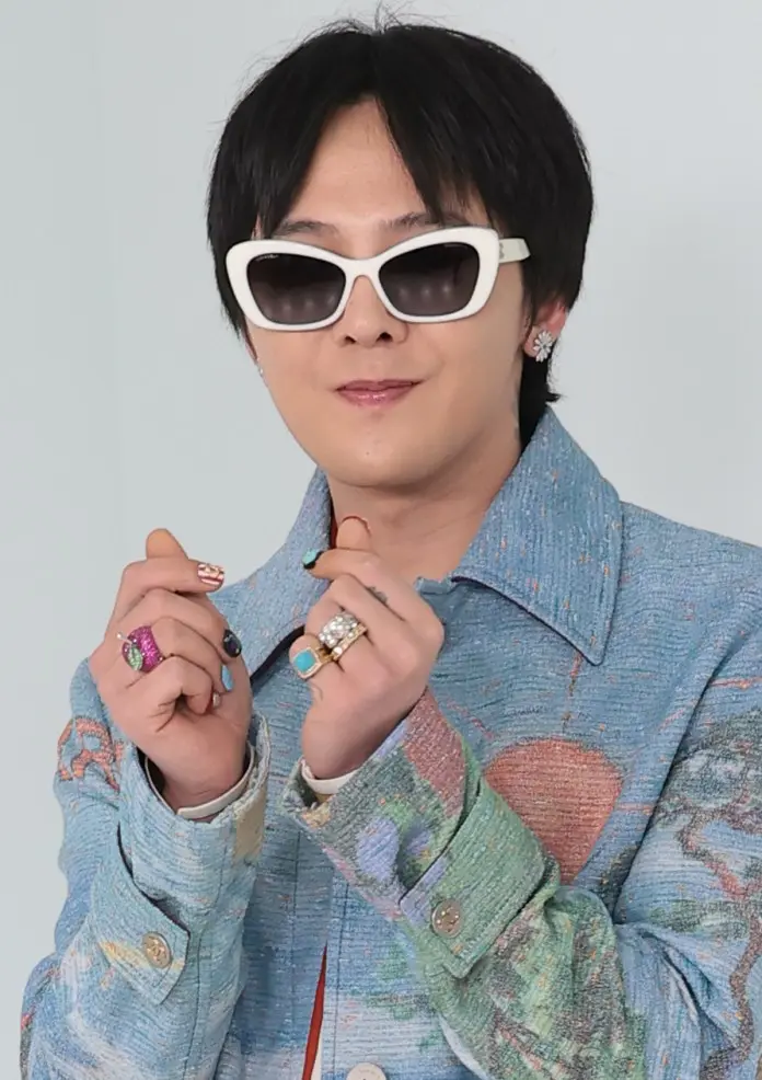 G-Dragon wearing stylish white sunglasses and a colorful jacket, showing jewelry on their hands.