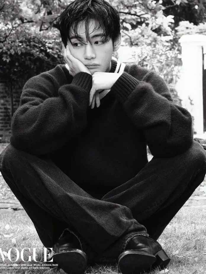 BTS V in a black sweater and dark jeans sits cross-legged on the grass, resting his chin on his hand with a thoughtful expression. The image is in black and white, featuring soft, natural surroundings.