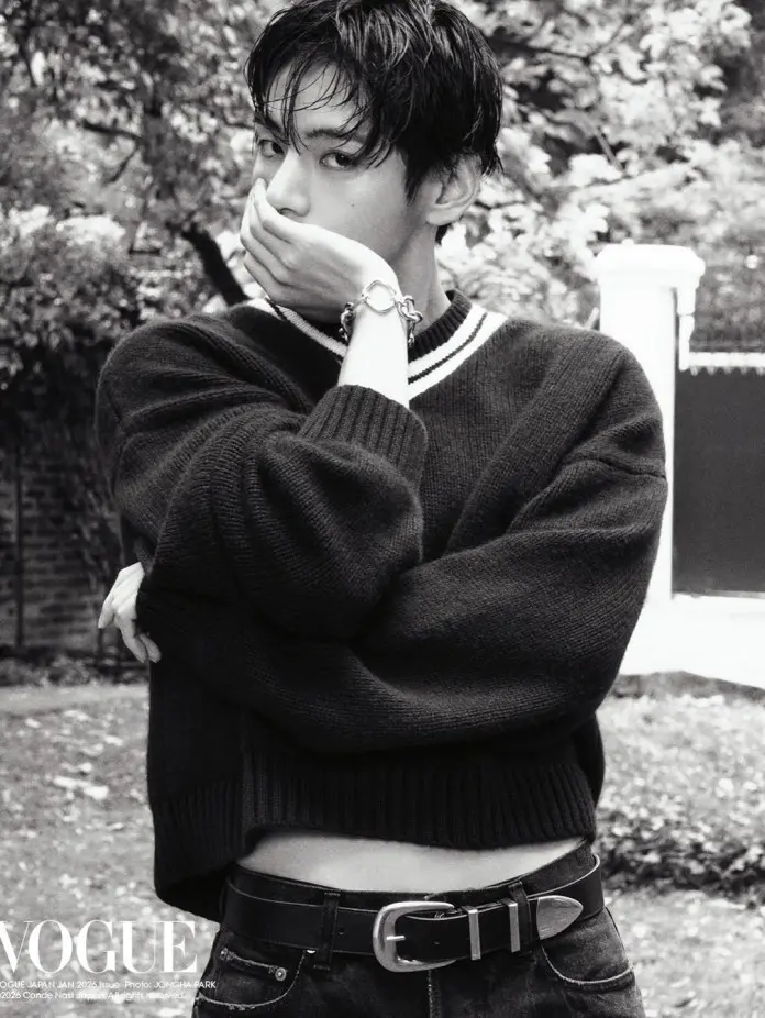 BTS V in a black and white image wearing a cropped black sweater with a white collar, holding their hand to their mouth in a thoughtful pose, standing outdoors with blurred greenery in the background.