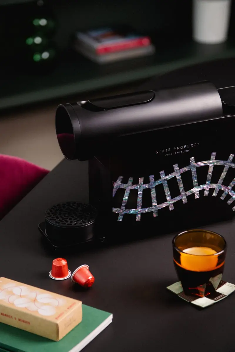 Black coffee machine with mother-of-pearl railroad-track inlay and State Property branding, beside red capsules and an espresso.