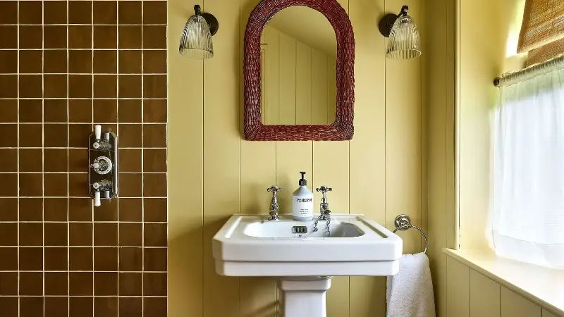 A yellow color-drenched bathroom with wall panelling, a white sink, an arched dark brown mirror, and two wall sconces. Caramel square wall tiles in the shower and a café curtain on the window.