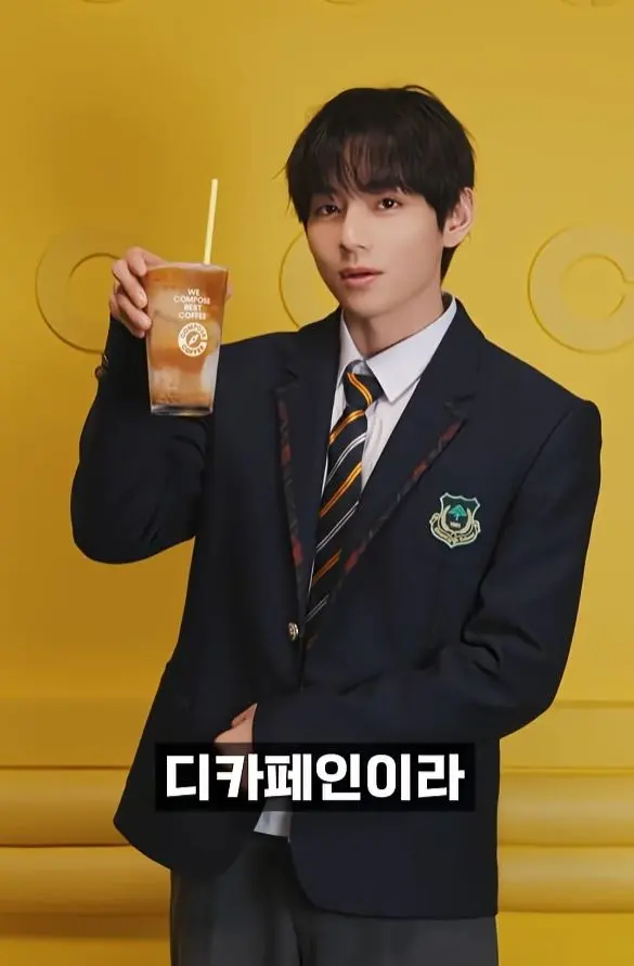 BTS V in a blazer holds a cup of iced coffee in front of a bright yellow background.