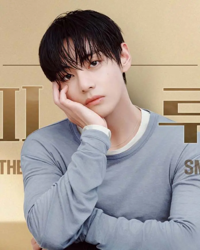 BTS V with wet hair, wearing a long-sleeved gray shirt, resting their chin on their hand, looking thoughtfully at the camera against a gold background.