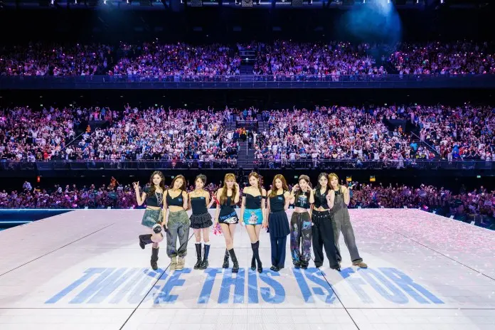 A South Korean girl group, TWICE, poses on stage in front of a cheering crowd during a concert, with the text 'TWICE THIS IS FOR' visible on the stage.